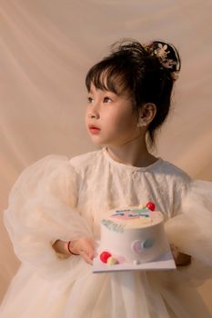 Adorable child in a white dress holding a cake, looking thoughtful indoors.