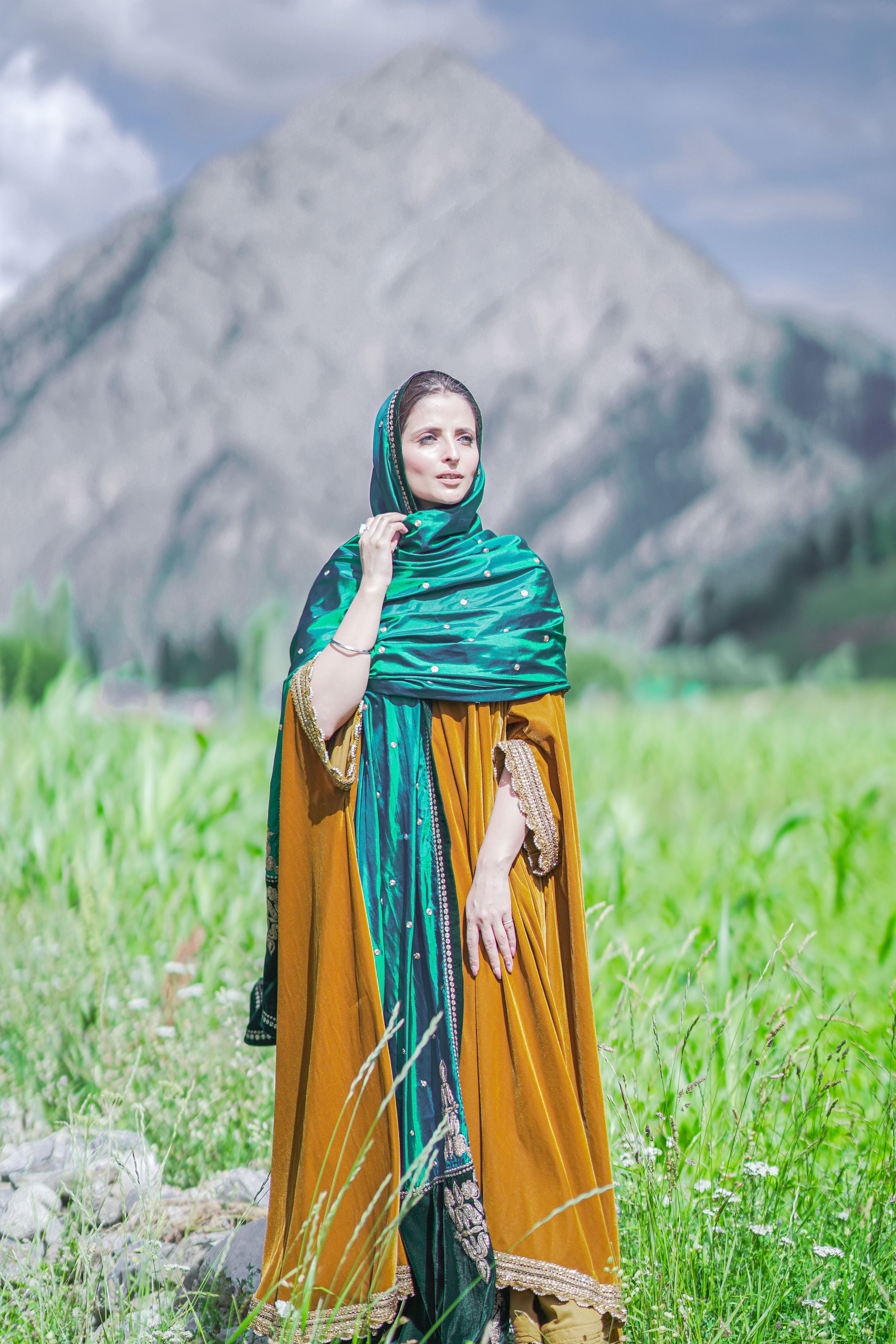 A woman in traditional attire stands outdoors with a majestic mountain backdrop.