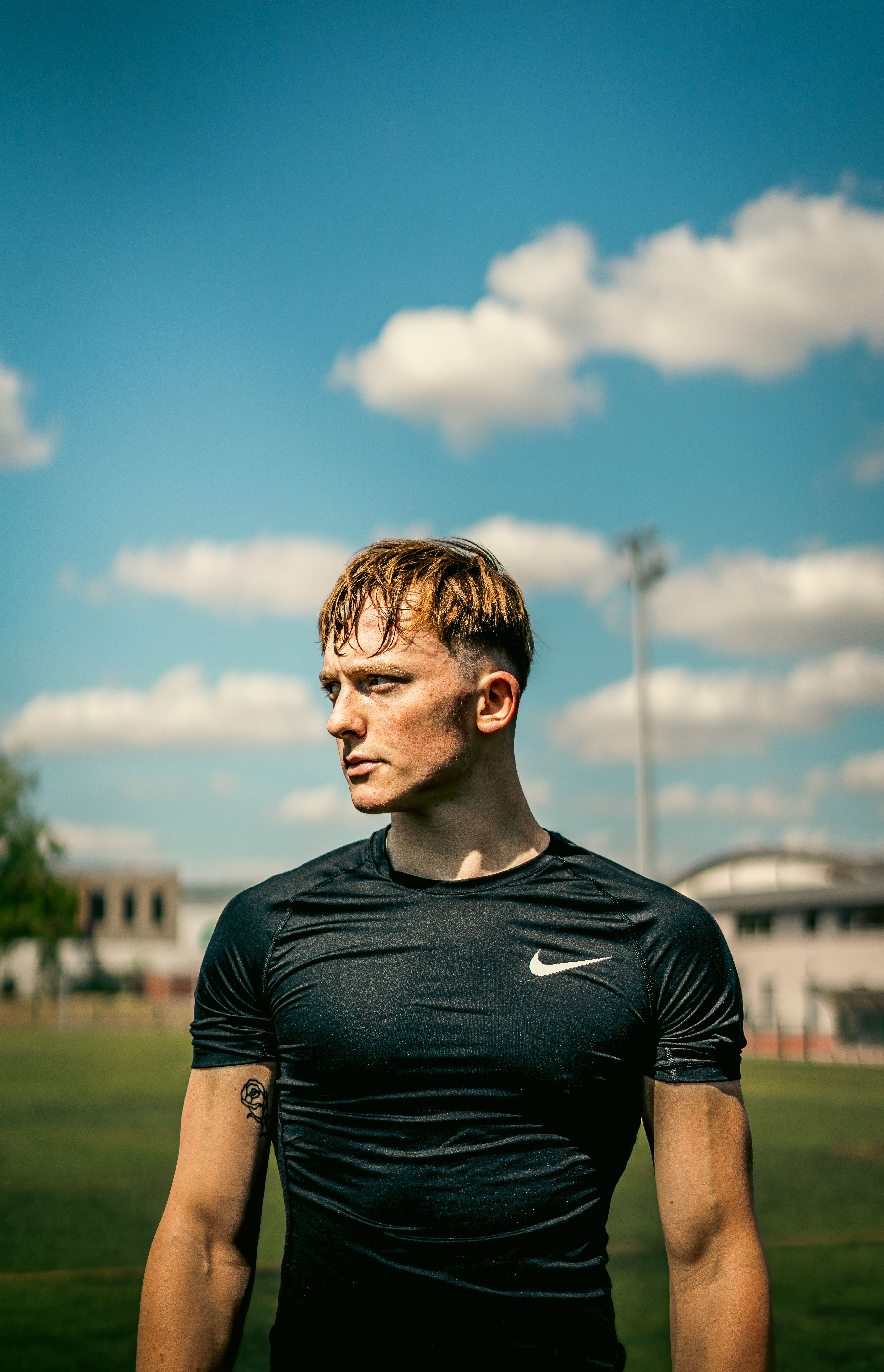 Fit young man in a black shirt outdoors with blue sky and clouds, embodying strength and fitness.