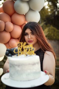 Woman holding a birthday cake with balloons in a festive outdoor setting.