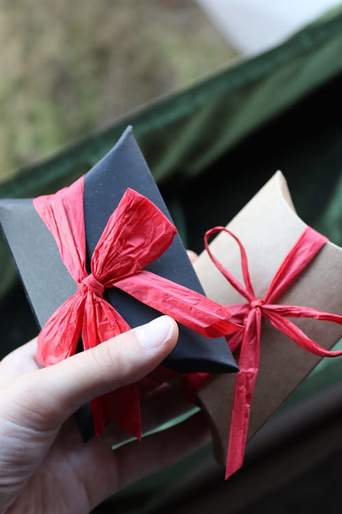 Hands holding two small gift boxes wrapped in red and brown paper with matching ribbons.