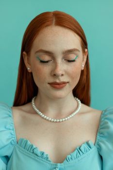 Portrait of a young redhead woman with freckles and pearls against a blue background.