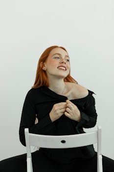 Portrait of a cheerful redhead woman in a black outfit sitting on a chair, posing playfully indoors.
