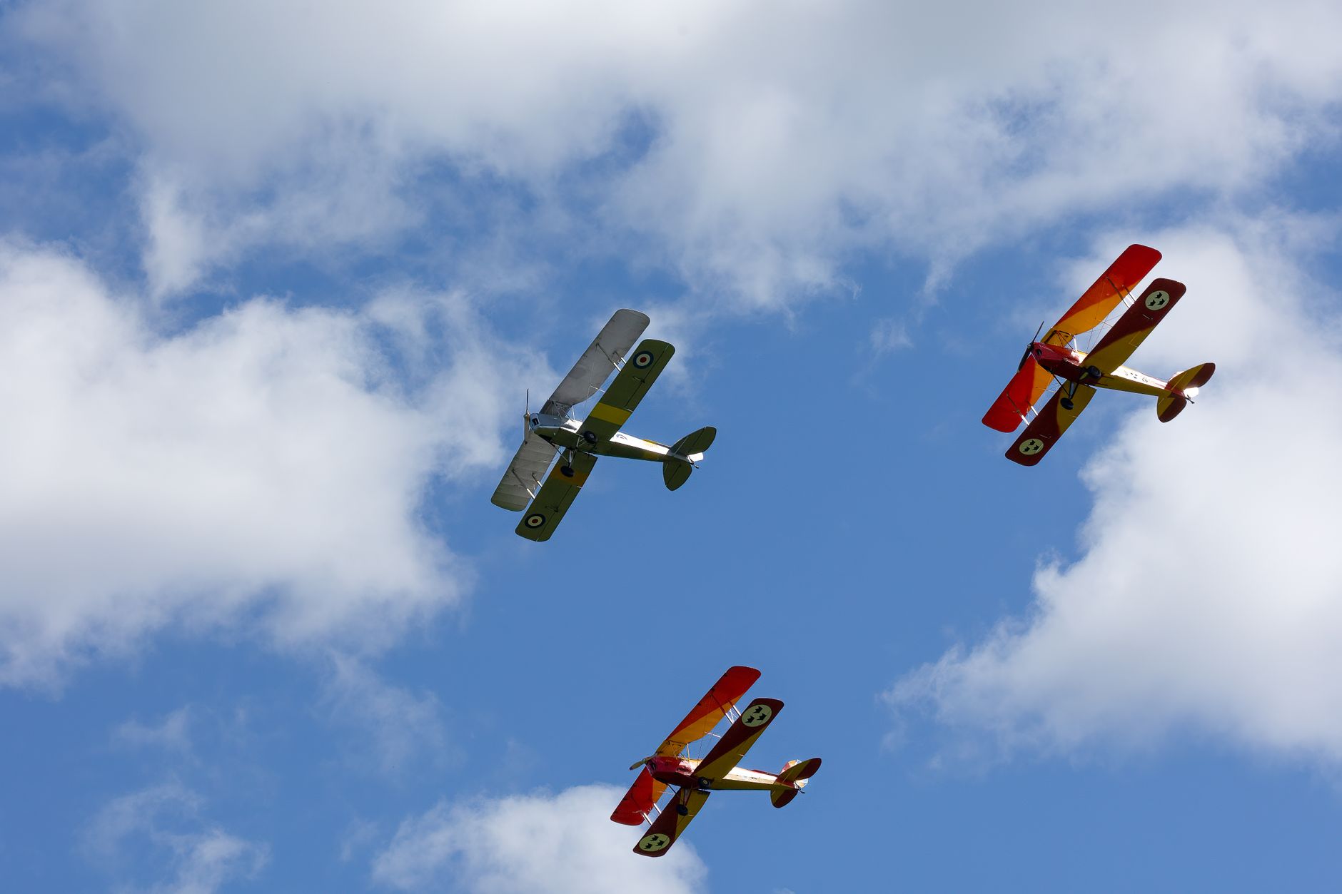 Three colorful vintage biplanes flying in formation against a vibrant blue sky with fluffy clouds.