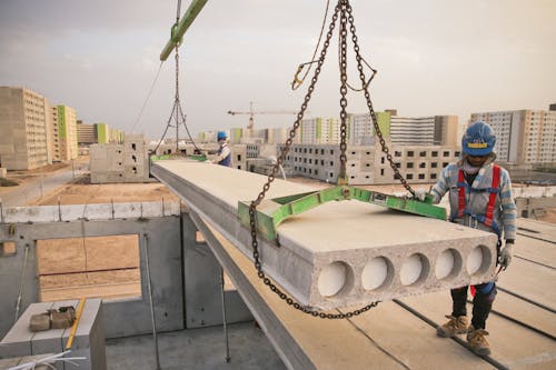 Anonymous workman in uniform and protective helmet standing near reinforced cement board hanging on metal chain of crane near modern multistory houses