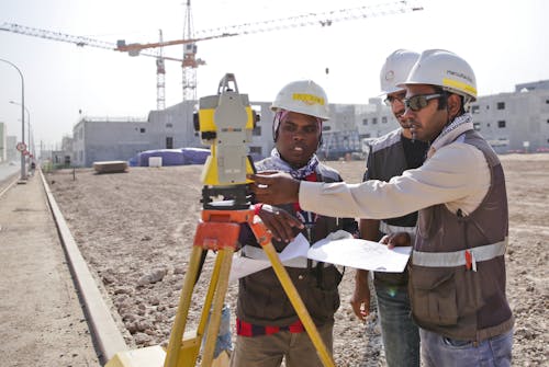 Surveyors in helmets using tachymeter on dry terrain near crane