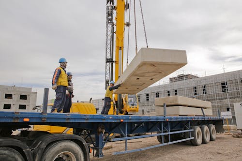 Group of unrecognizable male builders in protective hardhats standing on large chassis while crane putting reinforced cement layer on vehicle under cloudy sky