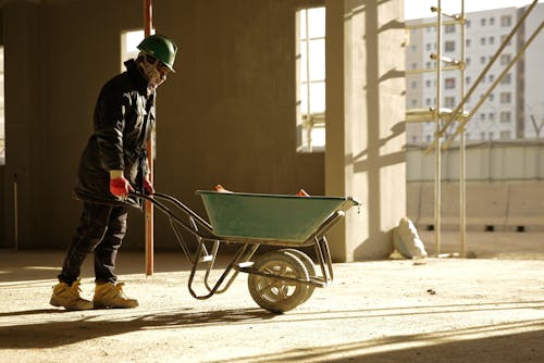 Side view of unrecognizable male employee in uniform and protective helmet carrying hand barrow with construction materials in contemporary building in sunlight