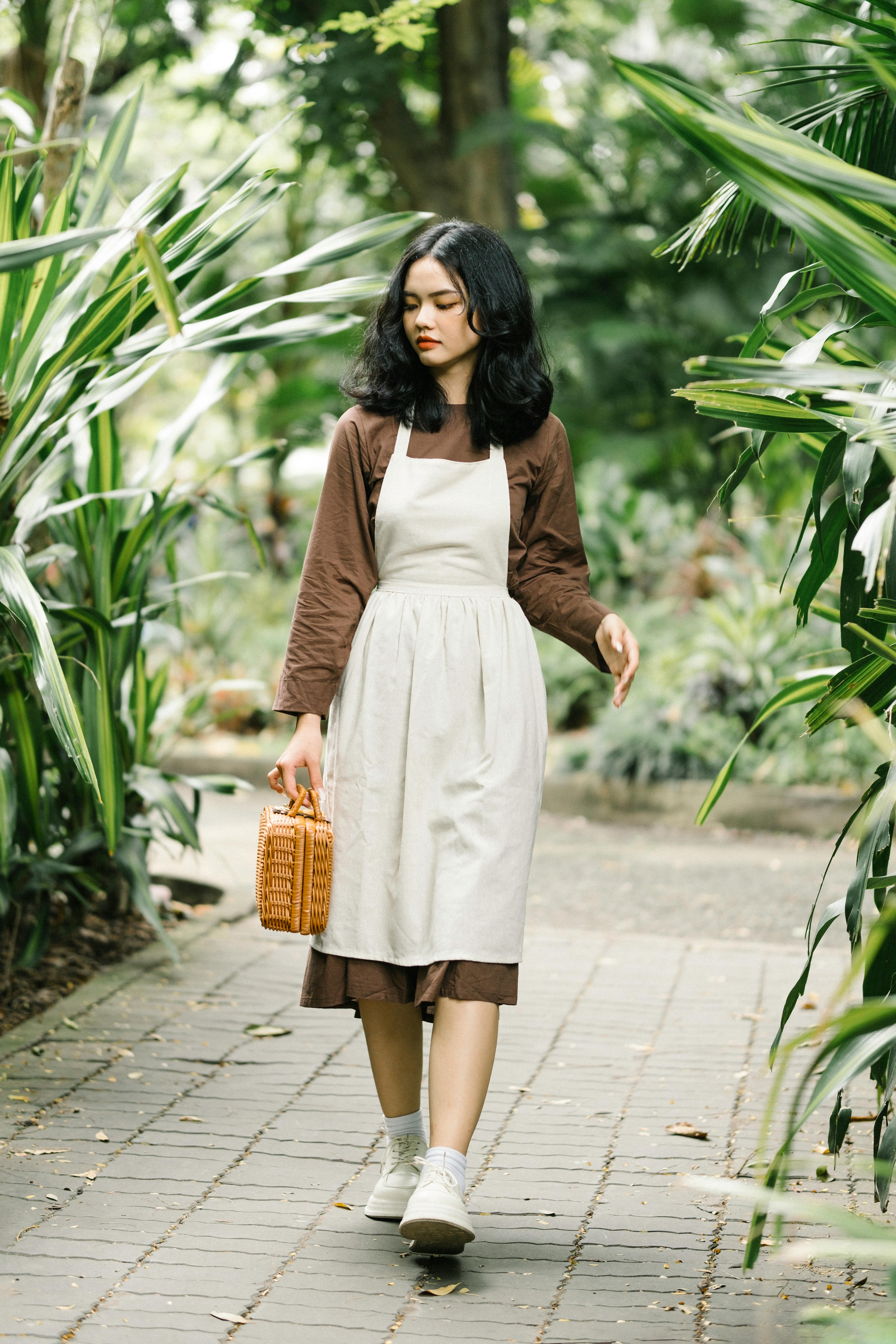 Fashionable young woman in apron dress walking through a lush garden holding a woven basket.