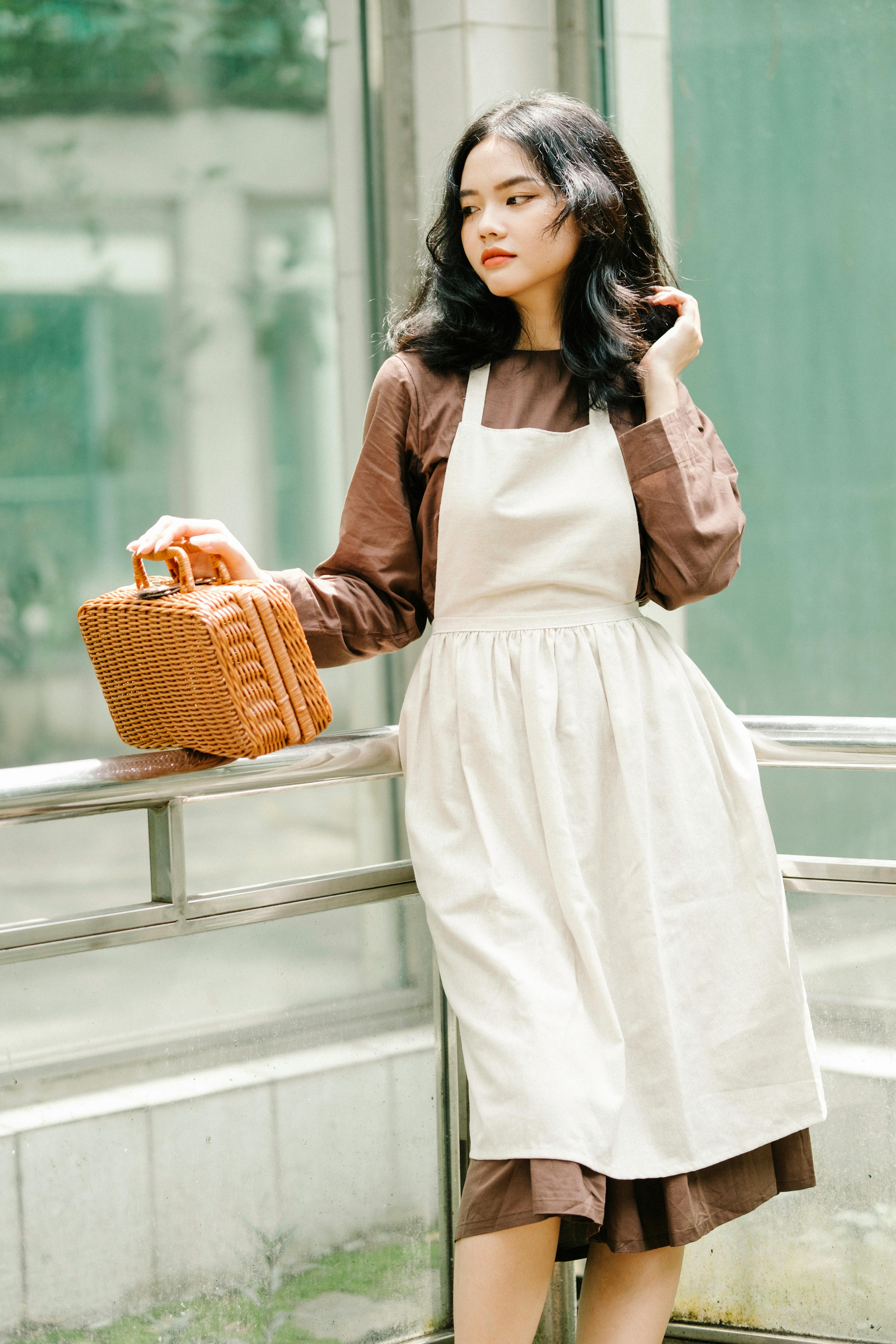 Stylish woman posing by a window in vintage attire holding a wicker bag.