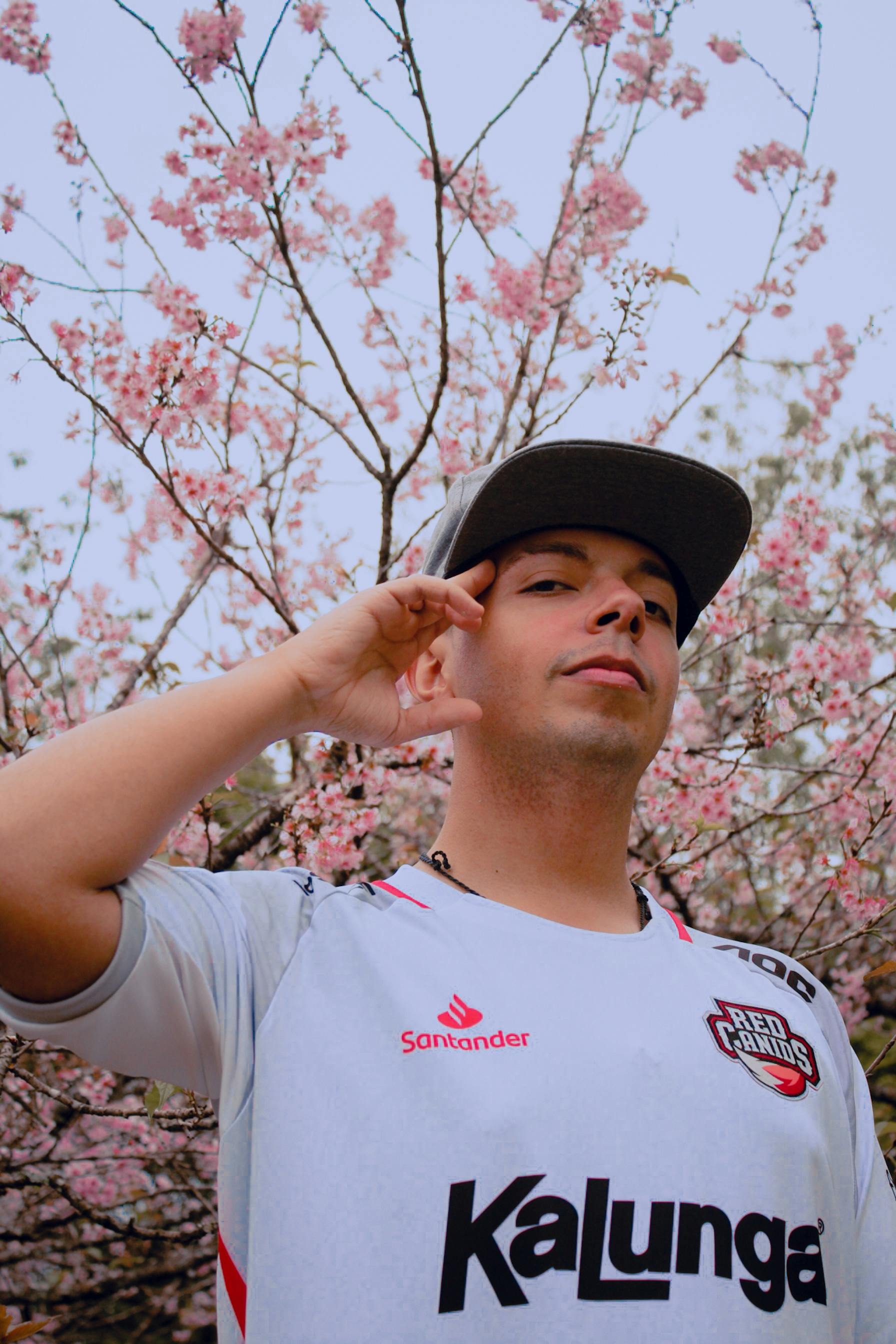 Young man in white t-shirt with black cap poses near blooming cherry blossoms.