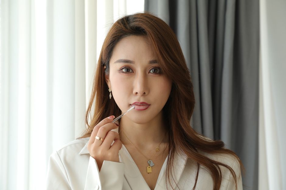 Asian woman applying lipstick in a softly lit indoor setting, exuding elegance.