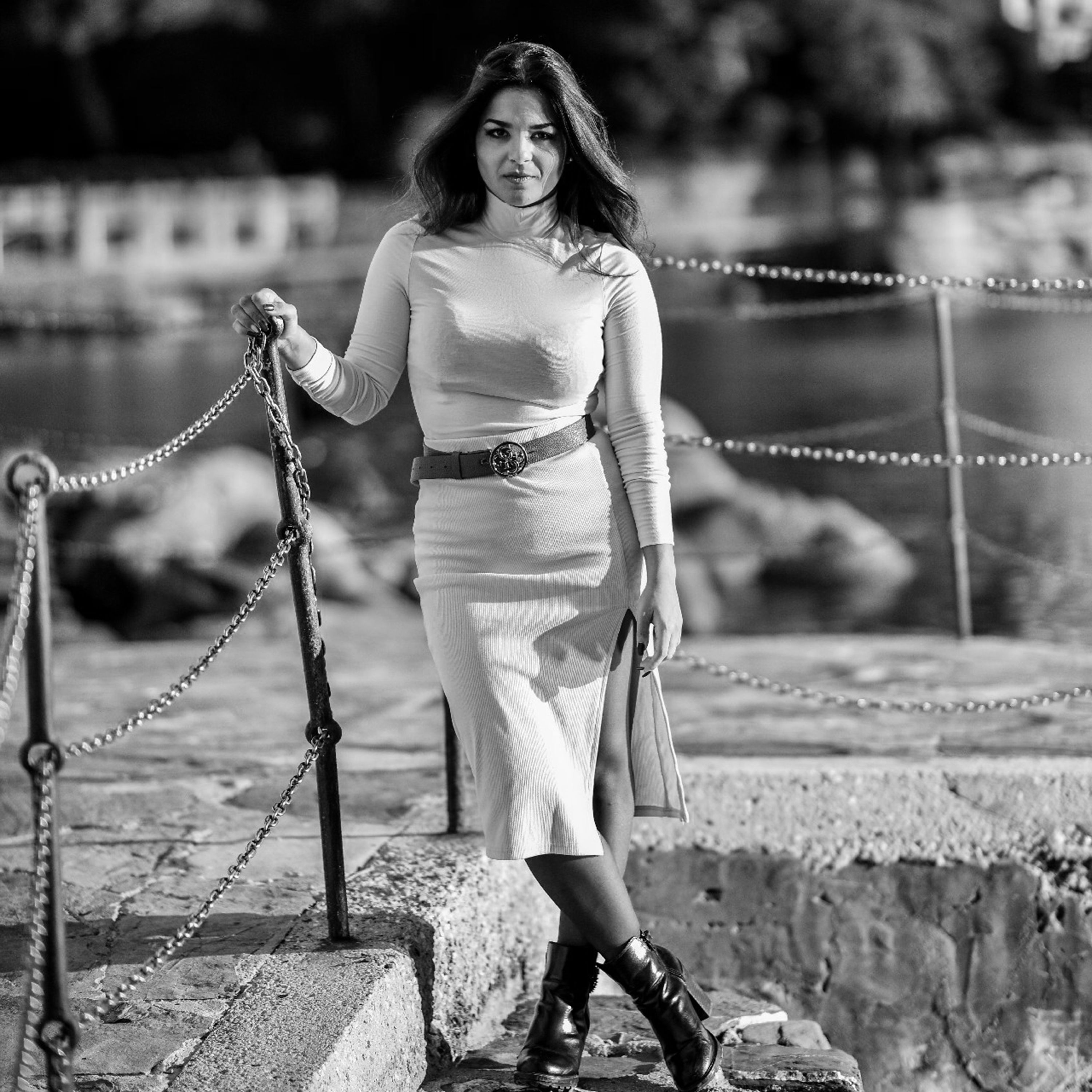 Elegant woman in white dress with black boots on concrete steps near water.