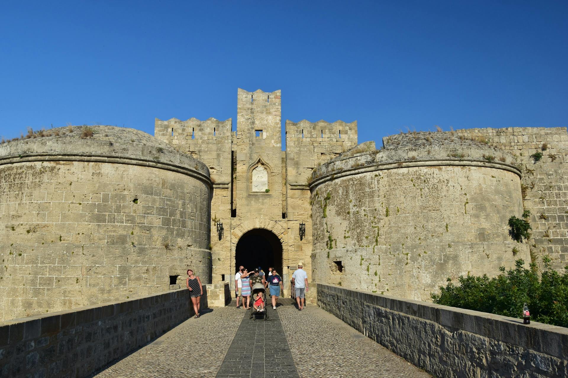 Tourists explore a historic fortress entrance in Rhodes, Greece under clear skies.