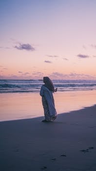 A person walks along a tranquil beach with a moody sunset sky, inspiring solitude and reflection.