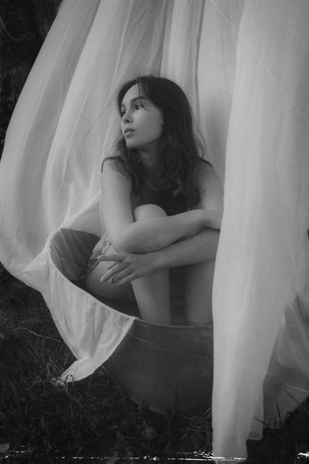 A contemplative woman sits wrapped in white fabric, captured in a grayscale portrait.