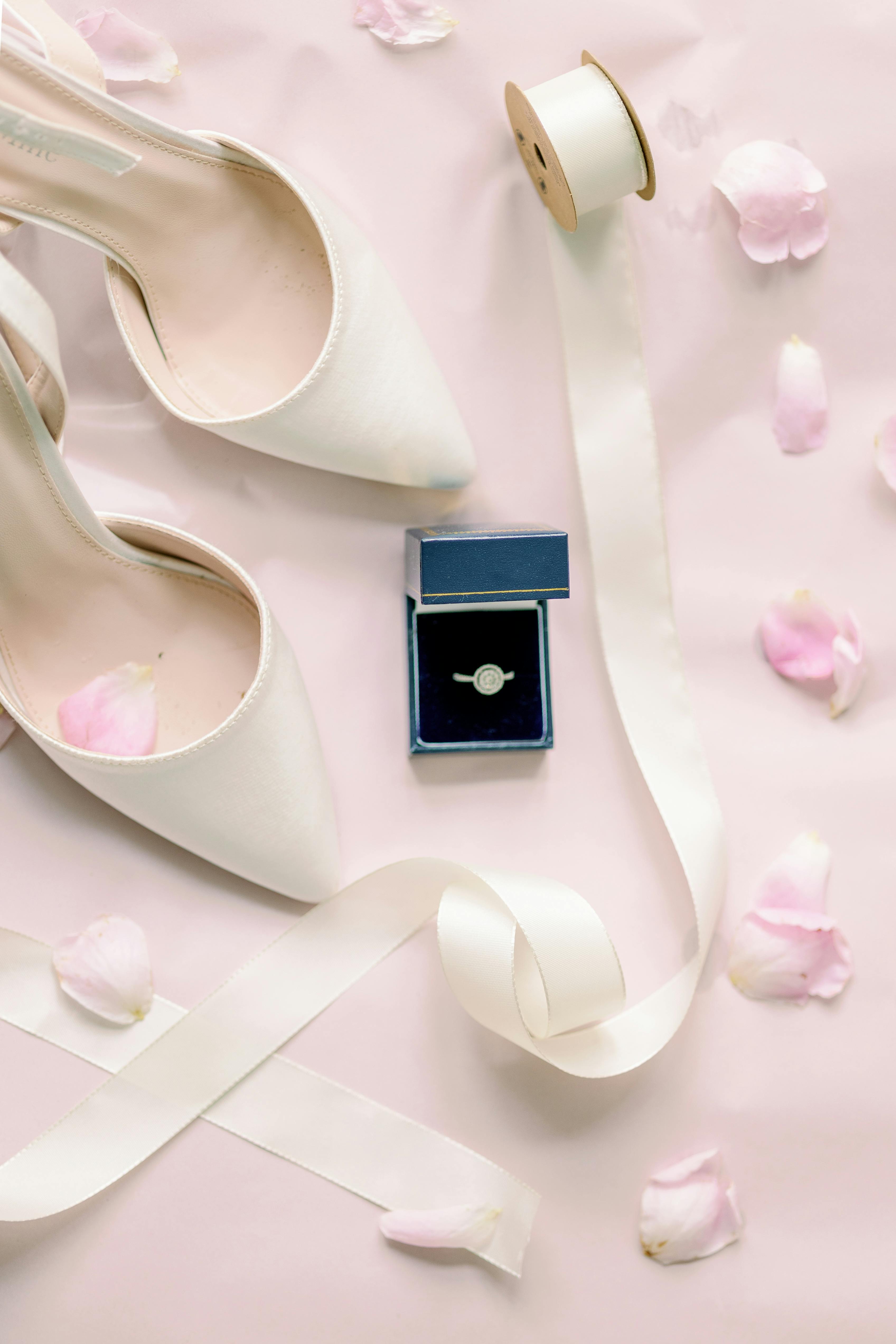 Flat lay of wedding shoes, ring, and ribbon with pink petals.