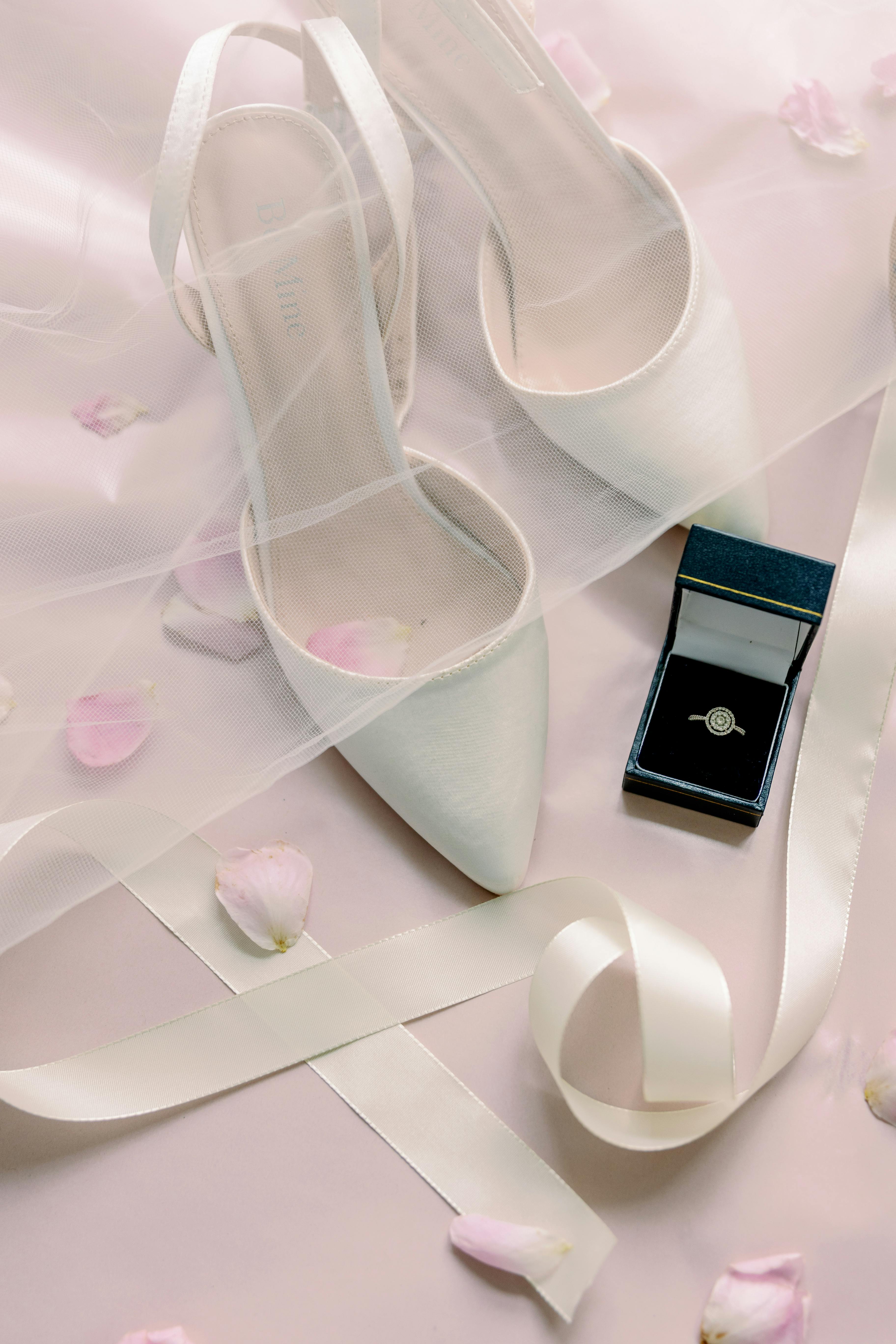 A close-up of wedding shoes, a ring in a box, and ribbon on a white background.