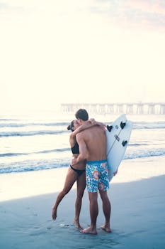 A couple embraces with a surfboard on Jacksonville Beach at sunset, capturing a moment of love.