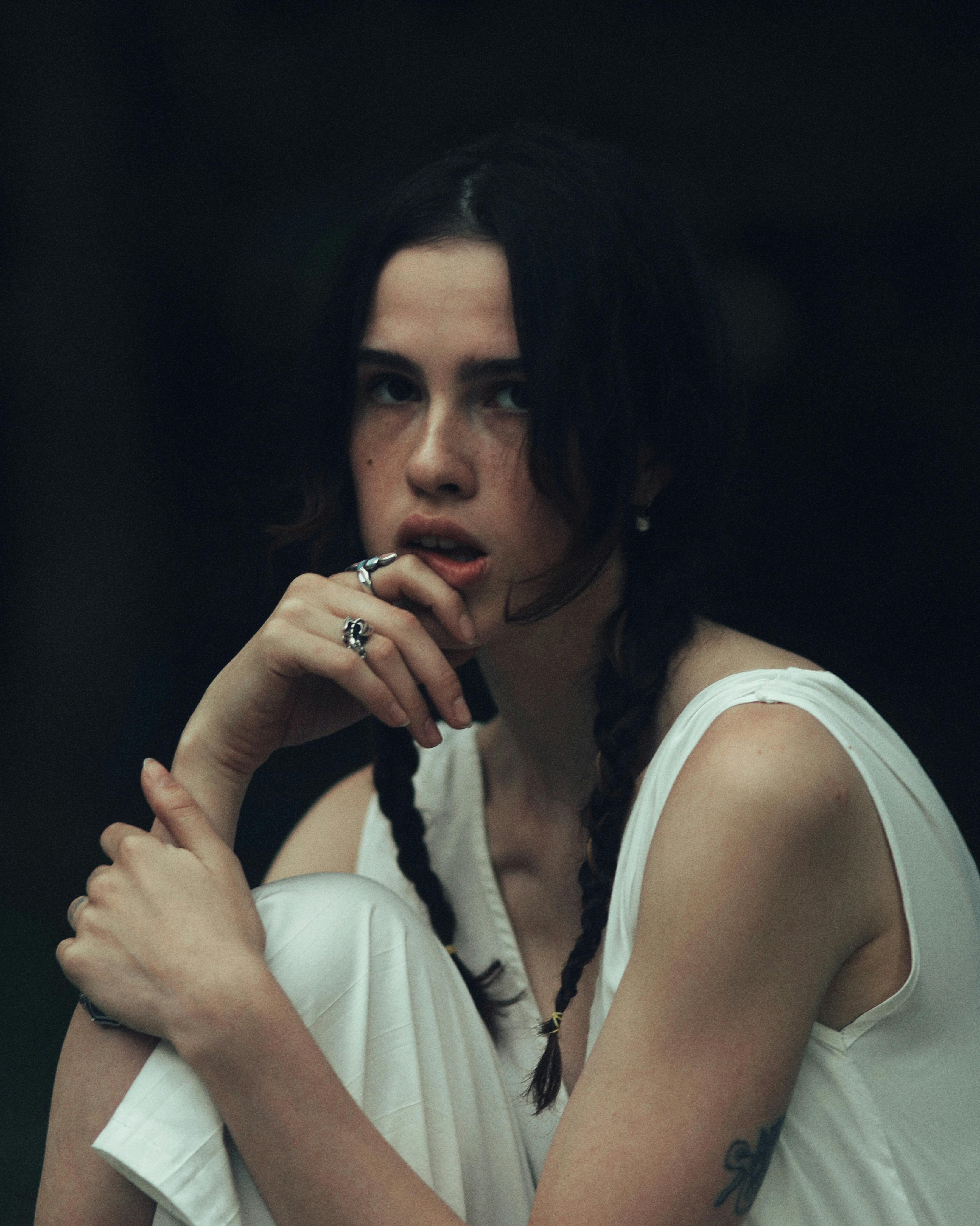 A young woman with braided hair sits in a casual pose, exuding a thoughtful and pensive mood.