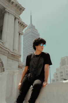 A young man with sunglasses poses stylishly against a city backdrop featuring iconic skyscrapers.