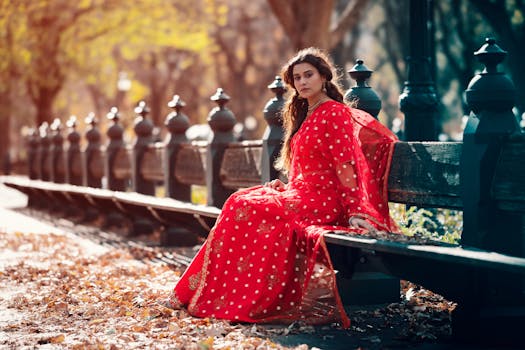 Woman in a vibrant red sari sits gracefully on a park bench amidst autumn foliage.