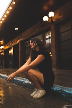 A young woman in a black dress sits on a curb under warm lights on a rainy city street.