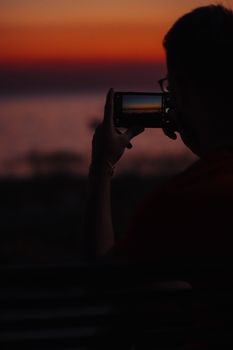 Person silhouetted taking a smartphone photo of a stunning sunset in Toroni, Greece.