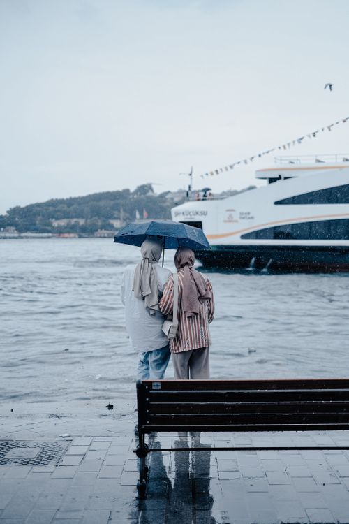 Free Couple standing under an umbrella by the sea on a rainy day, observing a cruise ship. Stock Photo