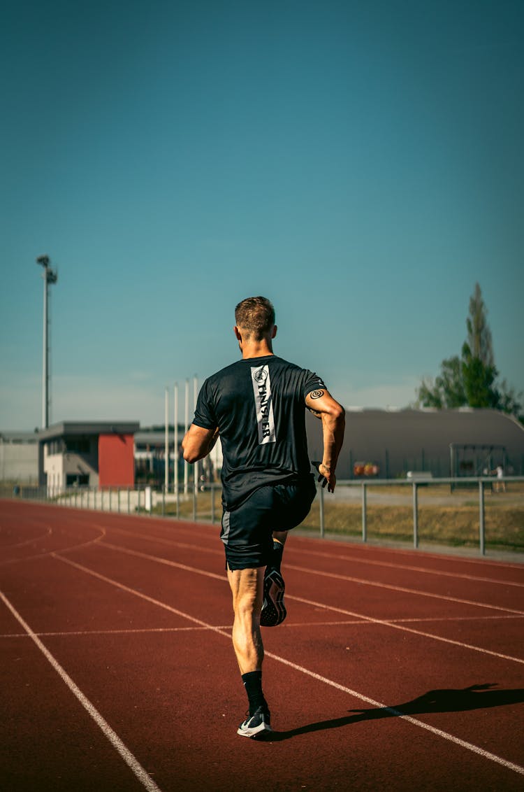 A Back View Of A Man In Black Shirt Running On The Track Field