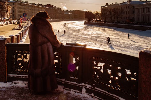 A person in a fur coat overlooking a frozen river in a snowy urban setting during winter.