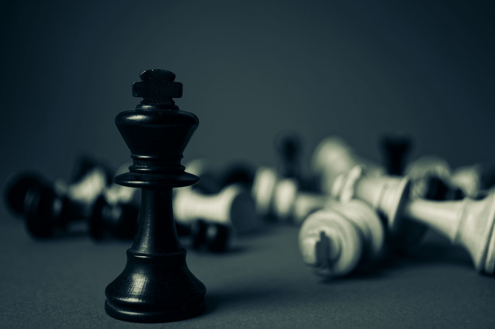 A black king chess piece standing upright in sharp focus while toppled white and black chess pieces lie scattered and blurred behind it
