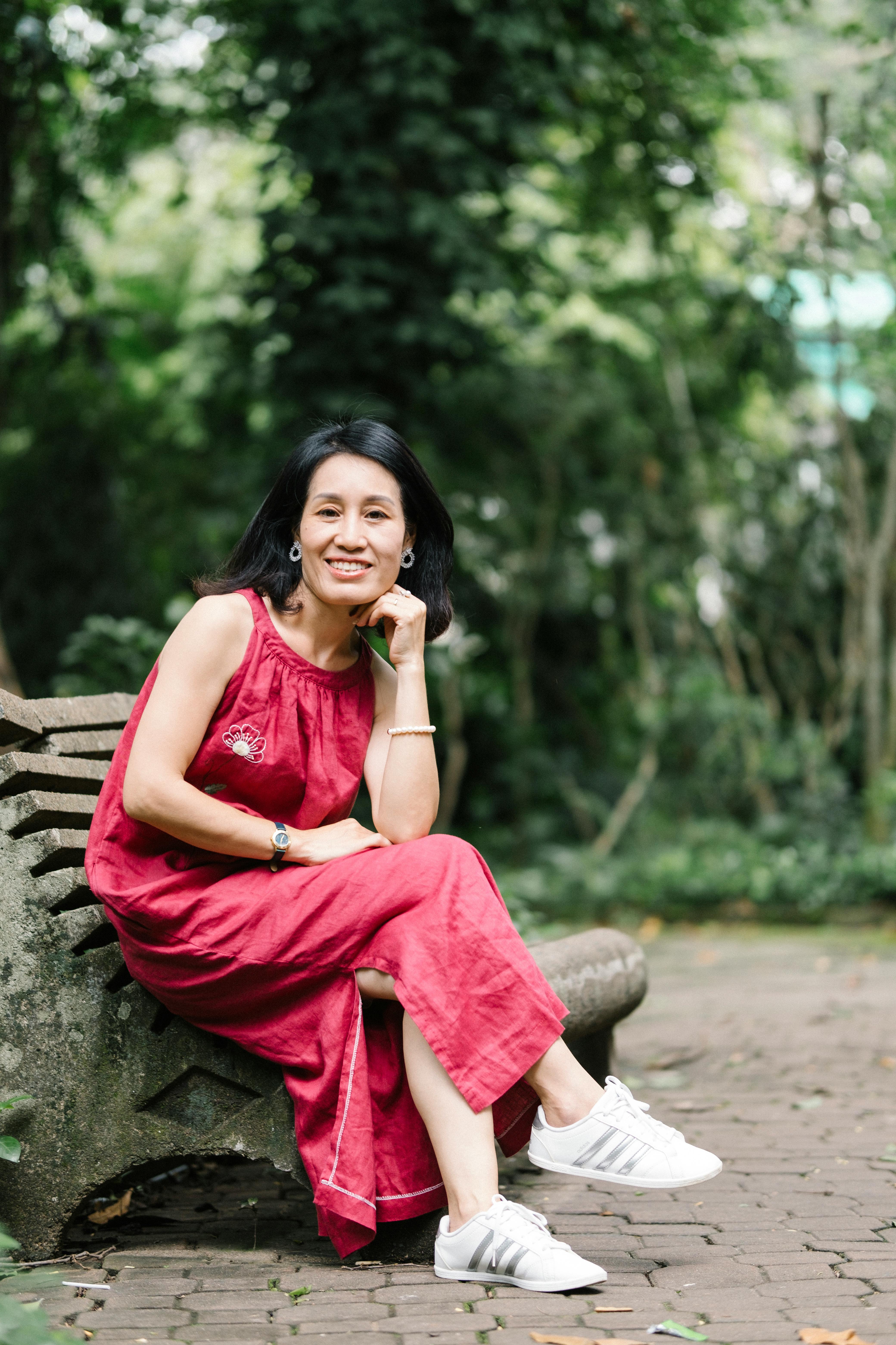 Asian woman in red dress sitting and smiling on a park bench surrounded by greenery.