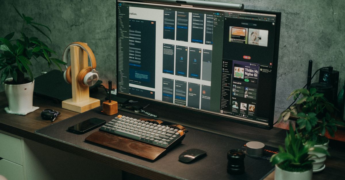 A sleek home office setup featuring a large monitor, keyboard, plants, and headphones on a desk.