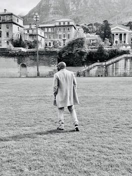A solitary figure walks across a grassy field with historic architecture and mountains in the background.