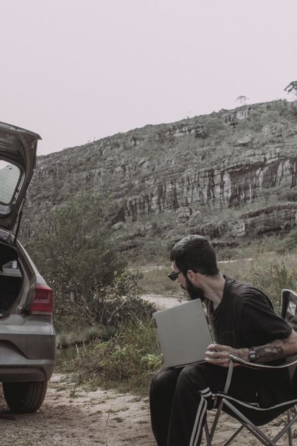 Man sitting on camping chair using laptop near car in scenic outdoor setting. Man sitting on camping chair using laptop near car in scenic outdoor setting.