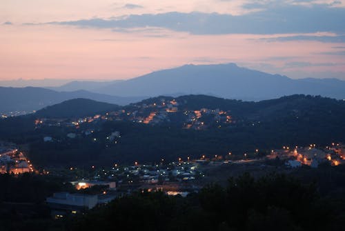 Wonderful scenery of illuminated with lights city build on highlands against mountain range and light pink cloudy evening sky