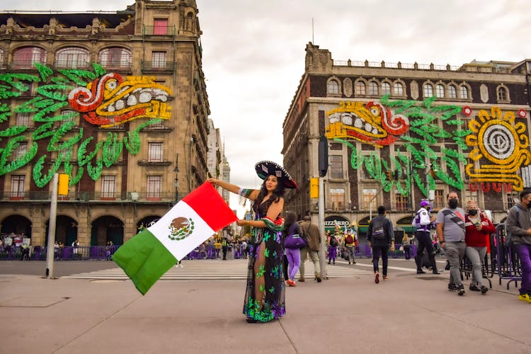 Woman In Mexican Costume And Hat Holding A Flag Around People Walking On Street