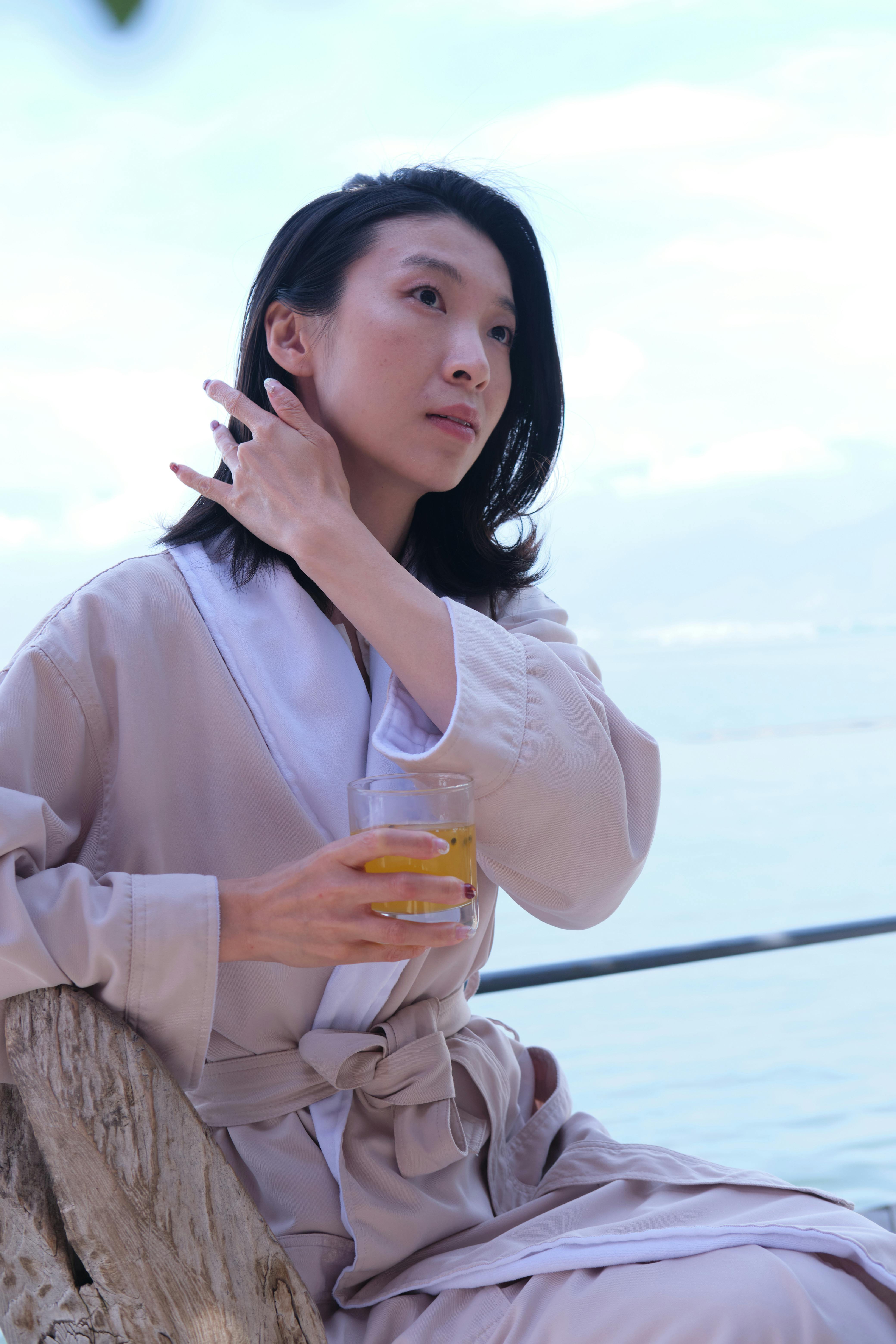 Free Serene woman in a robe enjoying a drink by the sea, embraced by tranquility and calm. Stock Photo
