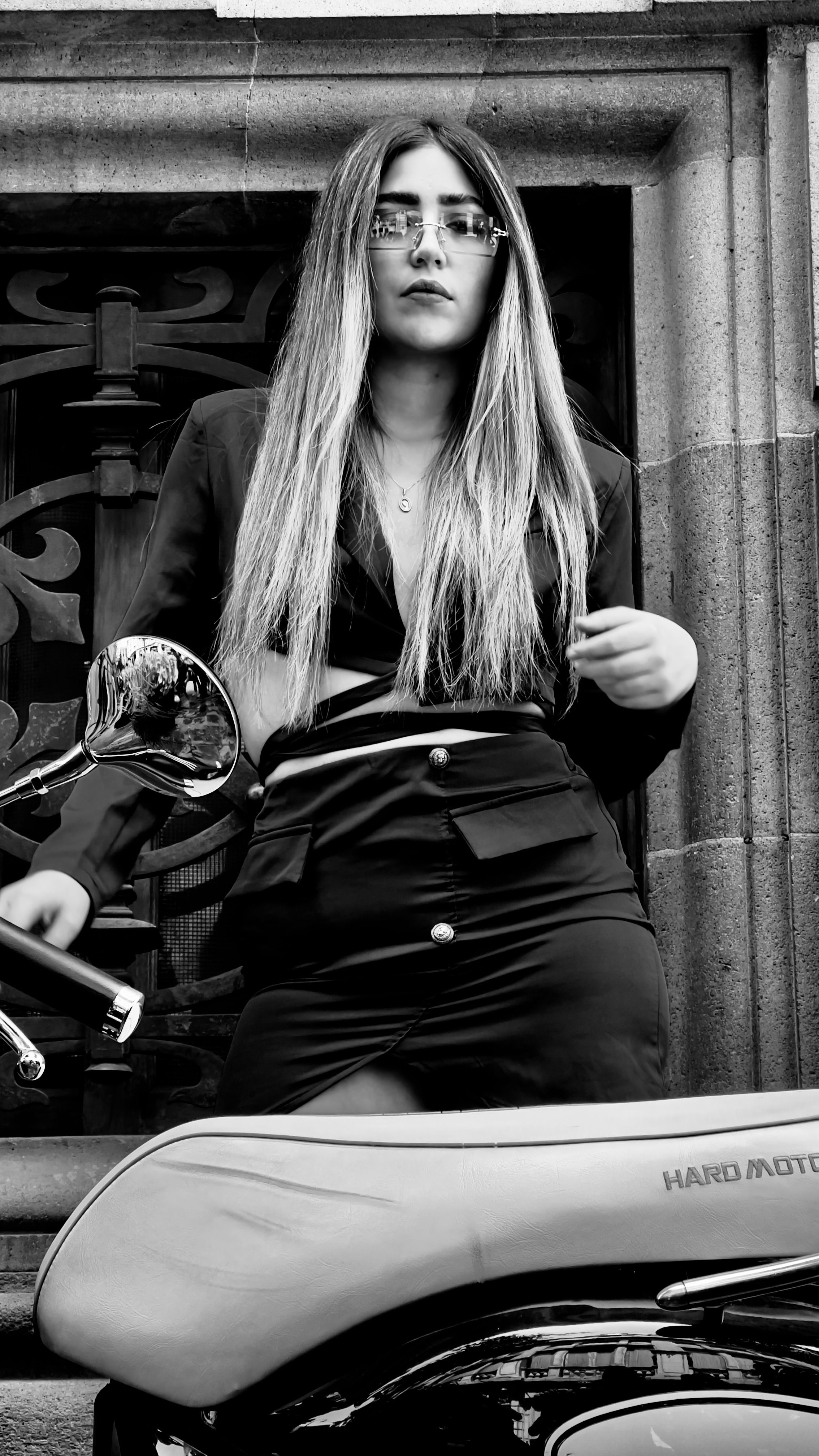 Monochrome image of a stylish woman standing next to a motorcycle, exuding retro fashion vibes.