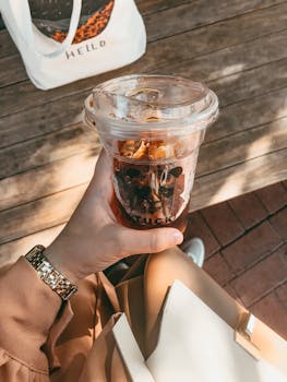 Close-up of a hand holding a plastic cup with iced coffee, outdoors on a sunny day.