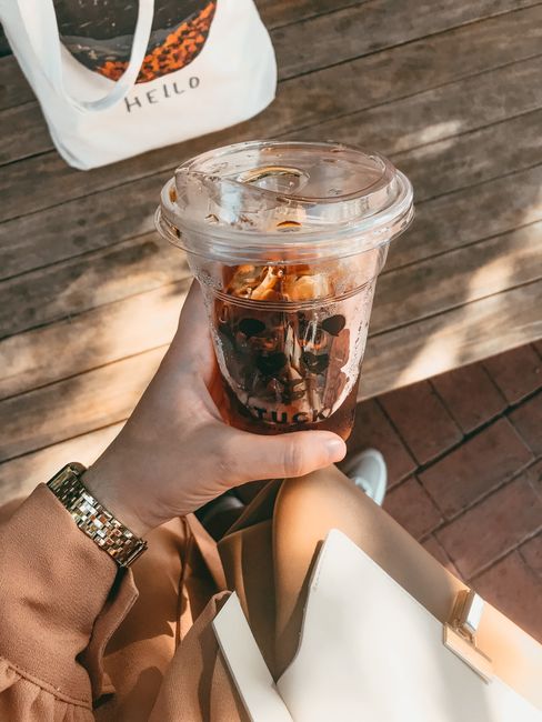 Close-up of a hand holding a plastic cup with iced coffee, outdoors on a sunny day.
