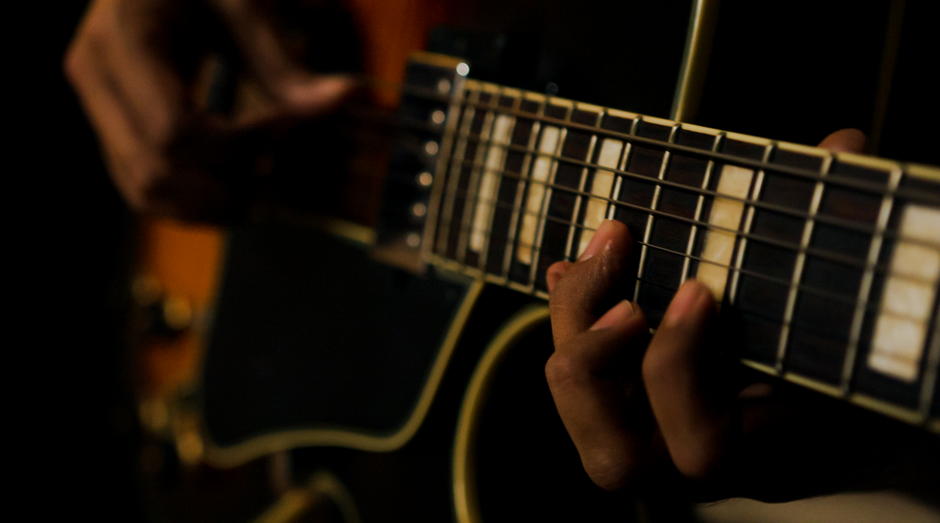 Close-up of a guitarist's hand fretting a chord on an electric guitar neck in low, warm light