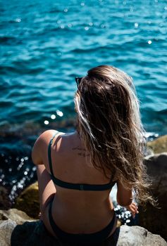 Back view of a woman in a bikini sitting on rocks by the sea under bright sunlight.