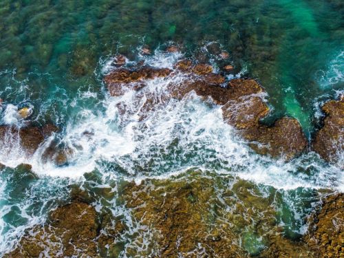Free Drone captures dynamic waves crashing on a rugged rocky shore, showcasing vibrant ocean hues. Stock Photo