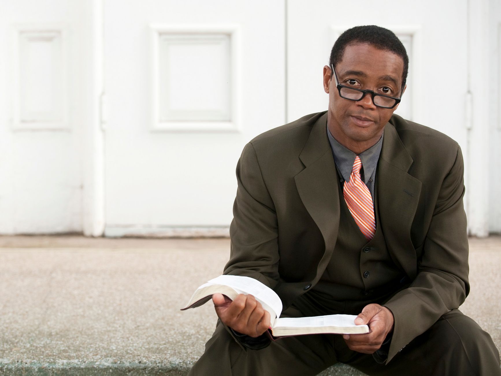 An African American man wearing a black suit and eyeglasses, holding a book while seated outdoors.