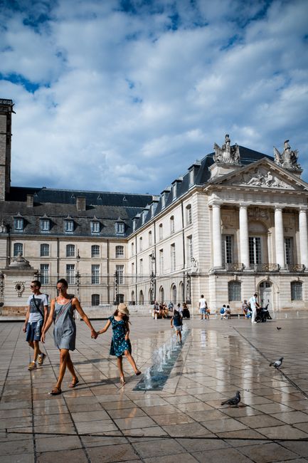 People enjoying a sunny day at a historic square with fountains and grand architecture.