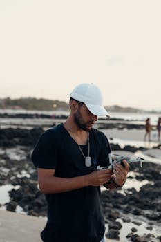 Young man with a drone on a rocky beach during sunset, wearing a cap and black shirt.
