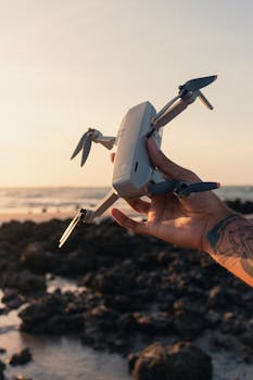 A tattooed hand holding a drone against a rocky beach at sunset, capturing the essence of adventure and exploration.