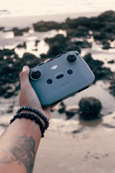 A tattooed arm holds a drone controller by the beach during sunset.